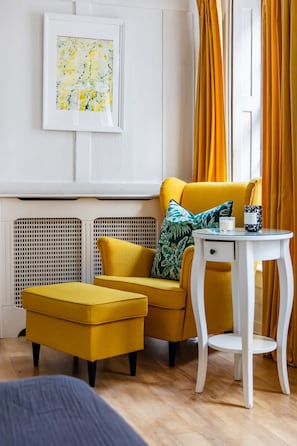 A room with a yellow sofa, a matching ottoman, a white side table with a candle, and a framed abstract painting on the wall. - Spacious 3BR 2BA Home in the Heart of Soho (London)
