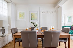A dining area with a wooden table, six chairs, and a vase of flowers. There are framed artworks on the wall and a green sofa in the background. - Spacious 3BR 2BA Home in the Heart of Soho (London)