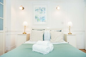 A neatly made bed with a green bedspread and two pillows, flanked by bedside tables with lamps, in a room with white walls and a framed artwork above the bed. - Spacious 3BR 2BA Home in the Heart of Soho (London)