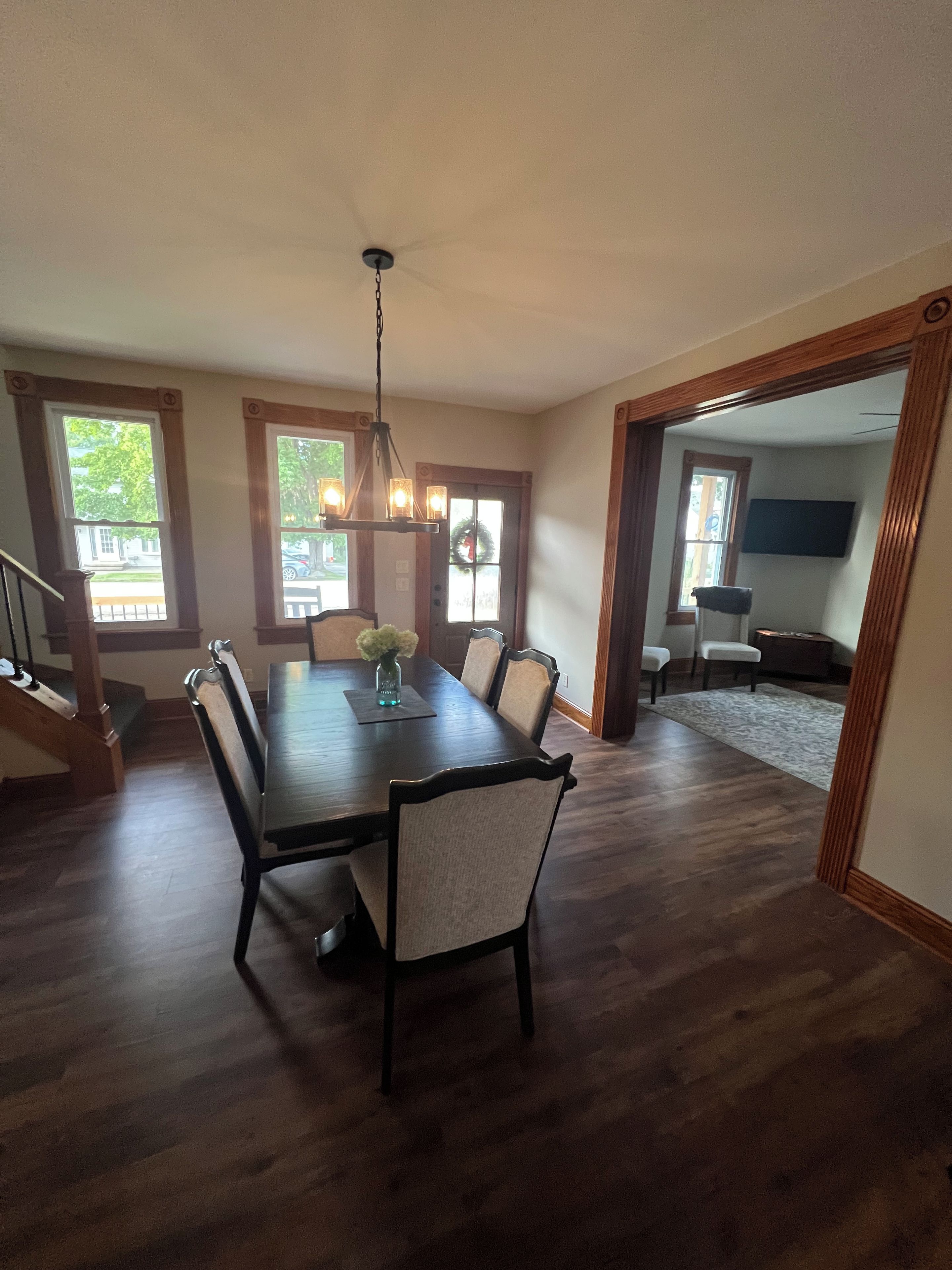 A dining area with a wooden table, chairs, and a chandelier. There is a staircase leading to another room.