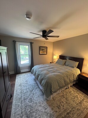 Ein Schlafzimmer mit einem Bett, Deckenventilator, einem Fenster mit Vorhängen, einem Nachttisch mit Lampe und einem gerahmten Bild an der Wand.
