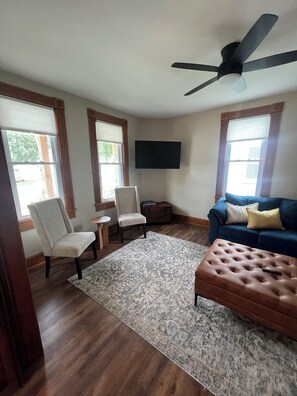 A living room with a ceiling fan, a blue sofa, a brown ottoman, and a grey patterned rug. - Beautifully Renovated Farmhouse next to Dorf Haus Supper Club (Sauk City)