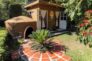 A small, single-story building with a covered porch, a door with curtains, and a garden with a palm tree and red flowers.