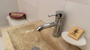 En handfat i badrummet med ett kromfärgat blandare, en tvålkopp och en toalett i bakgrunden.