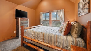 A bedroom with a sloped ceiling, a bed with patterned bedding, a television on a wooden dresser, and a window with curtains. - Mineral Mountain Lodge in Gilded Mountain (Lead)