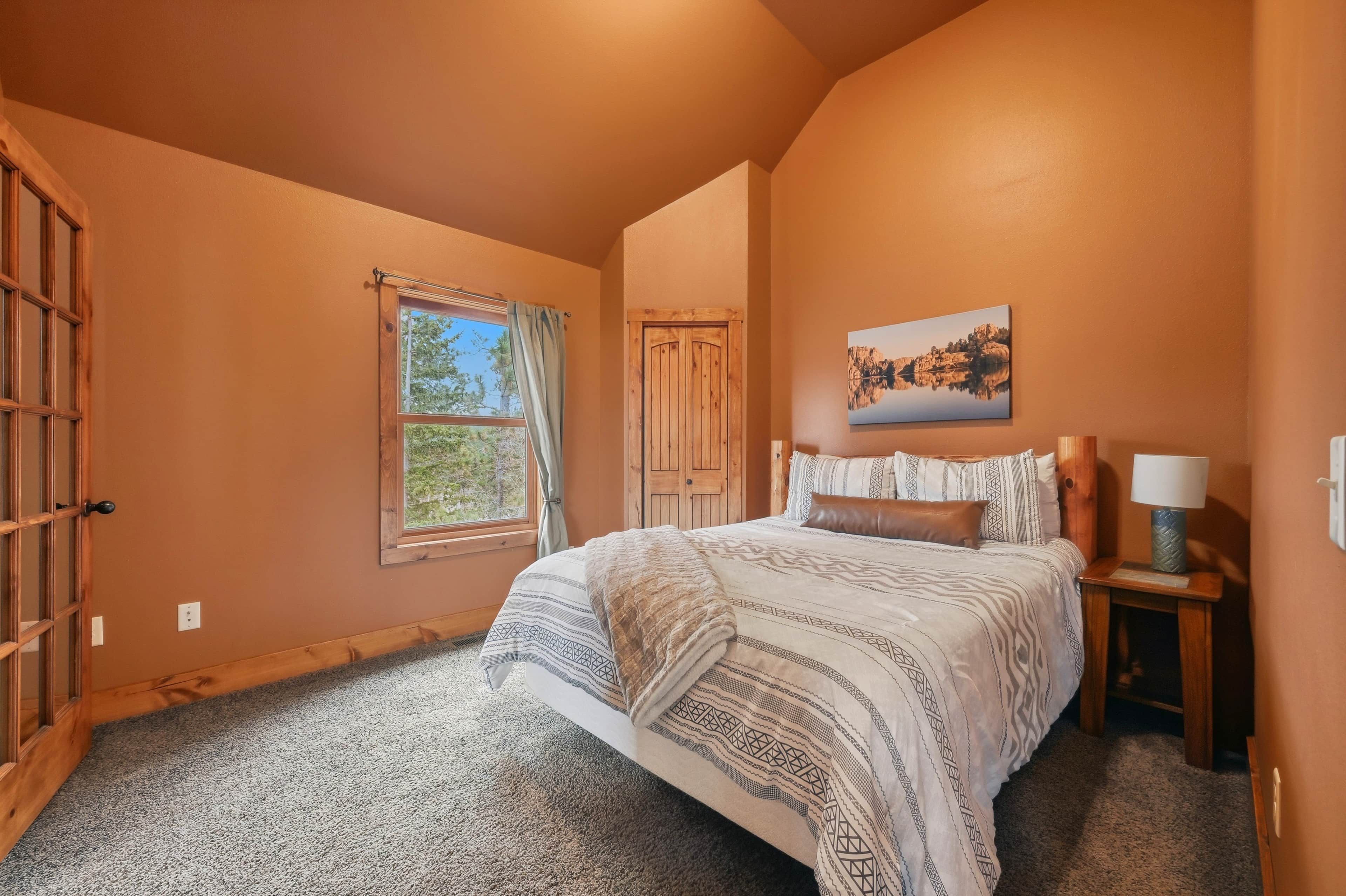 Quarto com uma cama grande, janela com cortinas, porta de madeira, quadro na parede e mesa de cabeceira com abajur.