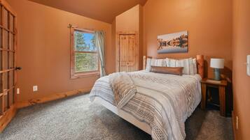 Quarto com uma cama grande, janela com cortinas, porta de madeira, quadro na parede e mesa de cabeceira com abajur.