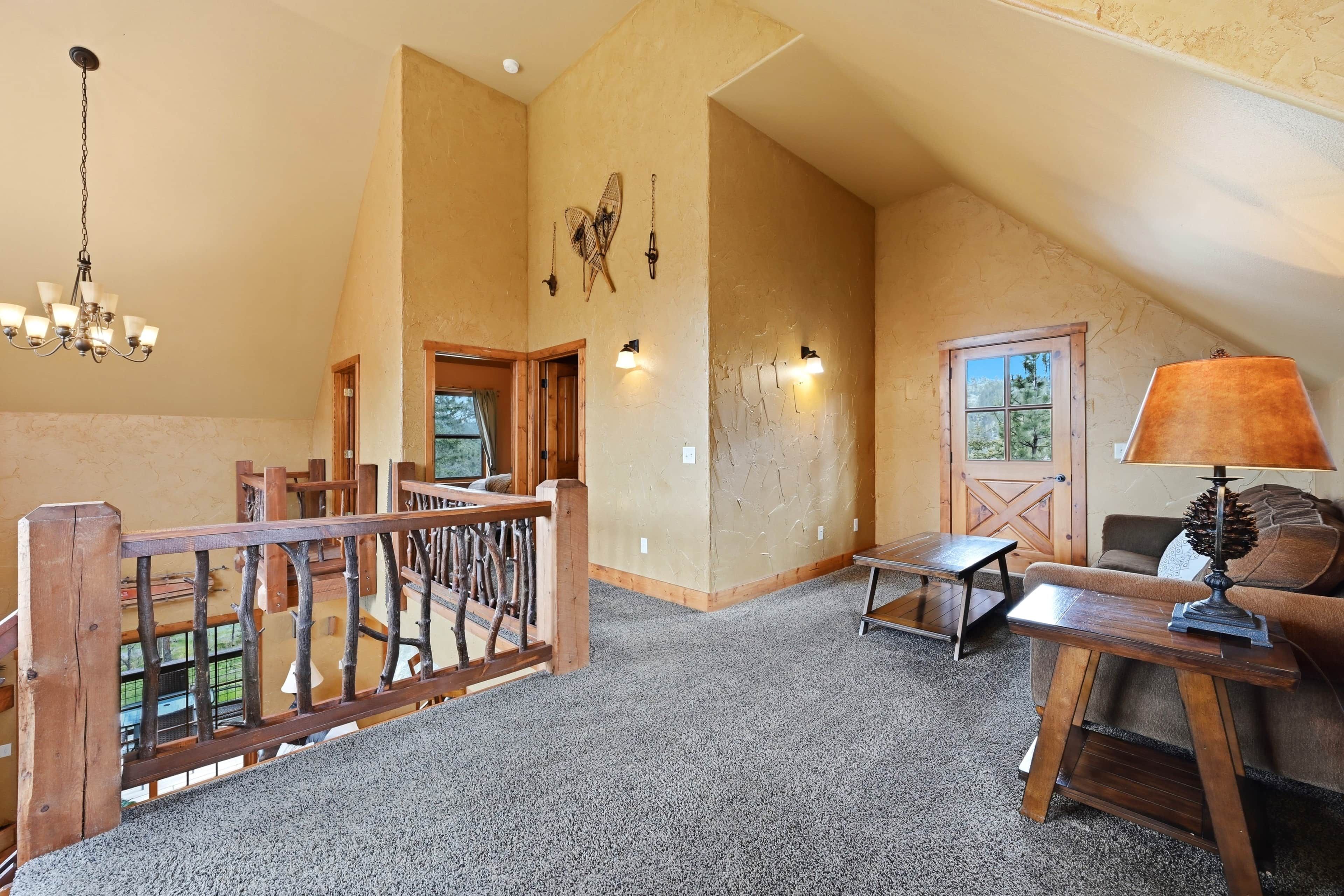A spacious loft with a high ceiling, a wooden staircase, a chandelier, a table with a lamp, and a view of the outdoors through a door.
