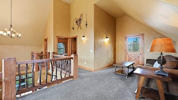 A spacious loft with a high ceiling, a wooden staircase, a chandelier, a table with a lamp, and a view of the outdoors through a door.
