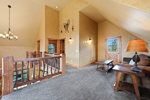 Un loft amplio con techo alto, una escalera de madera, un candelabro, una mesa con lámpara y vistas al exterior a través de una puerta.