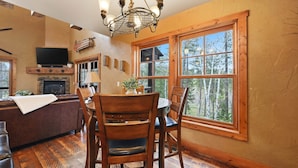 A dining area with a wooden table and chairs, a television mounted on the wall, and a large window with a view of trees. - Mineral Mountain Lodge in Gilded Mountain (Lead)