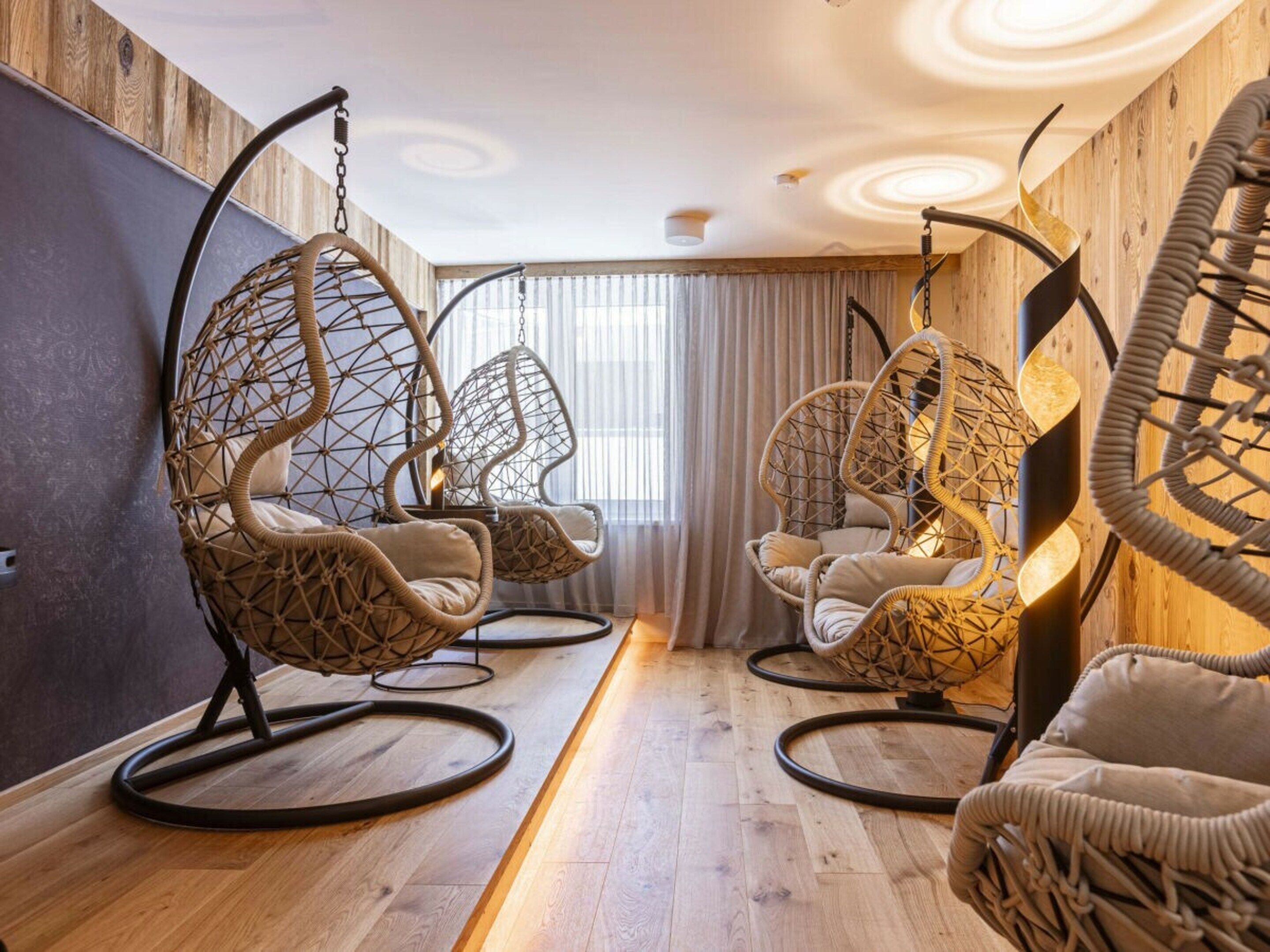 A room with hanging chairs, wooden flooring, and a large window with curtains.