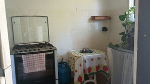 Una cocina con estufa a gas, una mesa con mantel estampado de frutas, un cilindro de gas azul y una planta en maceta.