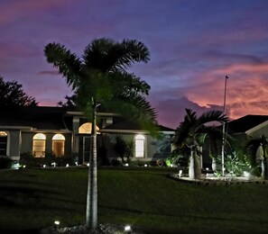 A residential property with a well-maintained lawn, a palm tree, and a house with arched windows and a lit entrance. - Beautiful home with heated saltwater pool in sought after Cape Coral, Florida. (Cape Coral)