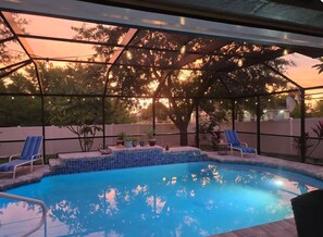 En pool med solstolar och krukväxter, omgiven av ett takförsett område med utsikt över träd och en solnedgång.