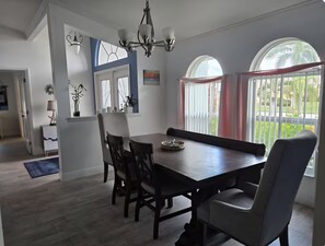 A dining area with a wooden table and chairs, a chandelier, and a view of greenery through large windows. - Beautiful home with heated saltwater pool in sought after Cape Coral, Florida. (Cape Coral)