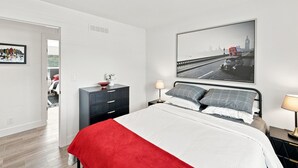 A bedroom with a bed, a dresser, a nightstand, a lamp, and a cityscape painting on the wall. - NEW Modern Contemporary West Side Retreat (Colorado Springs)