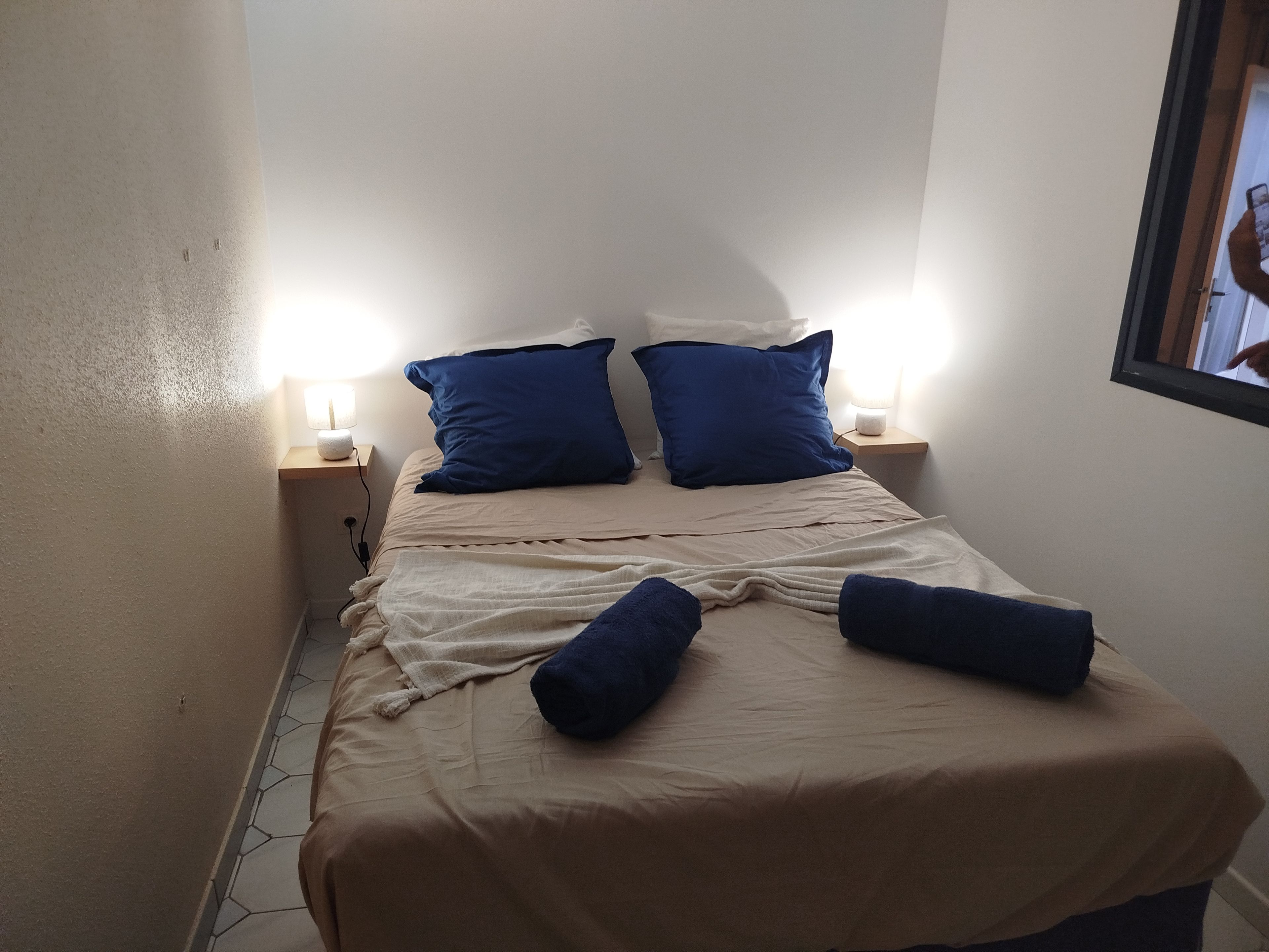 Un lit recouvert d’une couvre-lit beige et agrémenté de deux oreillers bleus, avec deux lampes de chevet placées de part et d’autre sur des tables de chevet en bois.