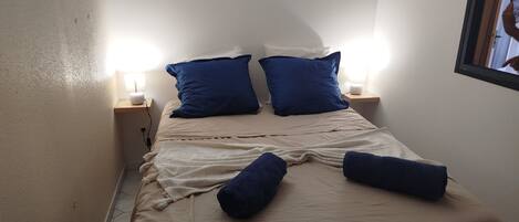 Un lit recouvert d’une couvre-lit beige et agrémenté de deux oreillers bleus, avec deux lampes de chevet placées de part et d’autre sur des tables de chevet en bois.