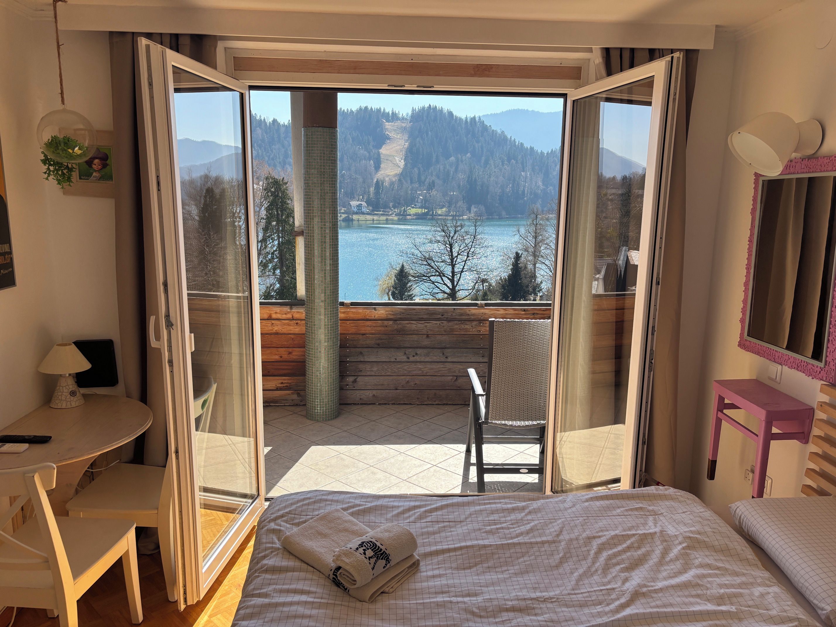 Quarto com cama, cadeira, escrivaninha e vista para um lago e floresta através de portas abertas.