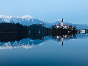 A serene lakeside view with a small island featuring a church and a red-roofed building, surrounded by snow-capped mountains. - Bled Castle & Lake View Apartment (Bled)