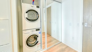 Une buanderie équipée de deux machines à laver superposées et d’un sèche-linge, ainsi que d’une conduite de ventilation et d’une porte donnant sur une autre pièce.