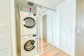 Une buanderie équipée de deux machines à laver superposées et d’un sèche-linge, ainsi que d’une conduite de ventilation et d’une porte donnant sur une autre pièce.
