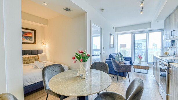 Une chambre d’hôtel moderne comprenant un lit, un coin repas avec une table ronde et des chaises, une cuisine et de grandes fenêtres offrant une vue sur la ville.