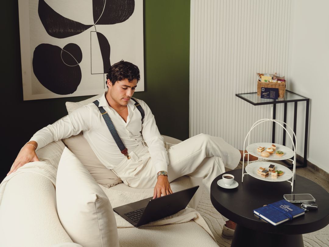 Eine Person sitzt auf einem Sofa mit einem Laptop, einem kleinen runden Tisch mit einem Buch und einer Tasse, und einem Tablett mit Essen und einem Getränk.