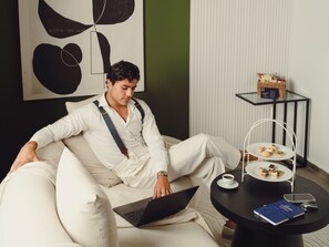 Una persona sentada en un sofá con una computadora portátil, una mesita redonda con un libro y una taza, y una bandeja con comida y bebida.