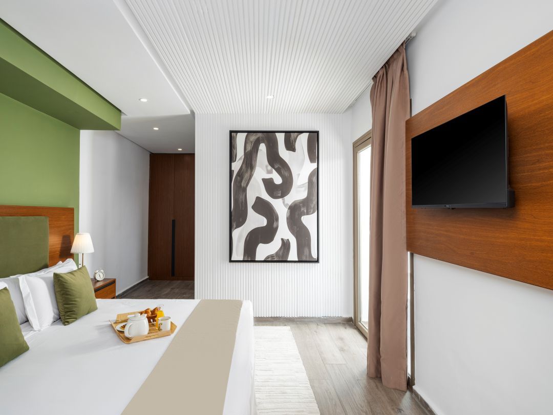A modern hotel room with a bed, a flat-screen TV mounted on the wall, a large abstract painting, and a wooden headboard.