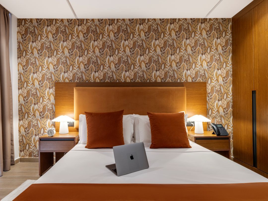 Ein Hotelzimmer mit einem großen Bett, zwei Nachttischen, einem Laptop, einem Telefon und einer gemusterten Tapete.