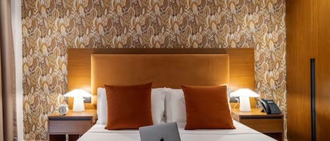 Habitación de hotel con una cama grande, dos mesitas de noche, una computadora portátil, un teléfono y un papel tapiz estampado.
