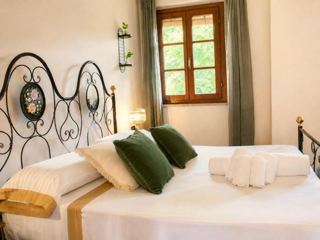 A neatly made bed with pillows and towels, a window with curtains, and a wall-mounted shelf with plants.