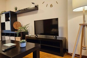 A modern living room with a flat-screen TV, a dark wooden coffee table, a remote control, a potted plant, and a wall-mounted shelf with decorative items.