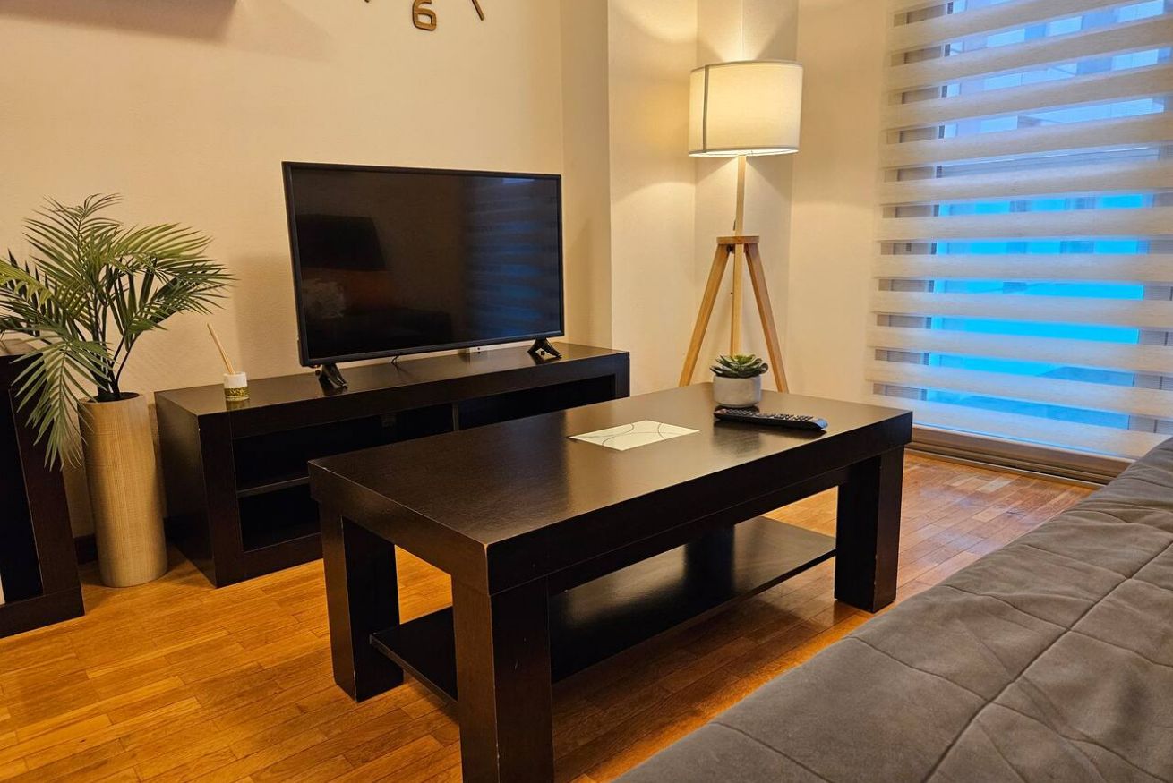 Ein modernes Wohnzimmer mit Fernseher, Couchtisch aus dunklem Holz, Stehlampe und einer Zimmerpflanze.
