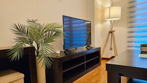 Een moderne woonkamer met een flatscreen televisie op een donker houten dressoir, een vloerlamp en een grote kamerplant.