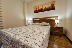 A hotel room with a bed, bedside lamps, a cityscape painting, and a wooden headboard.