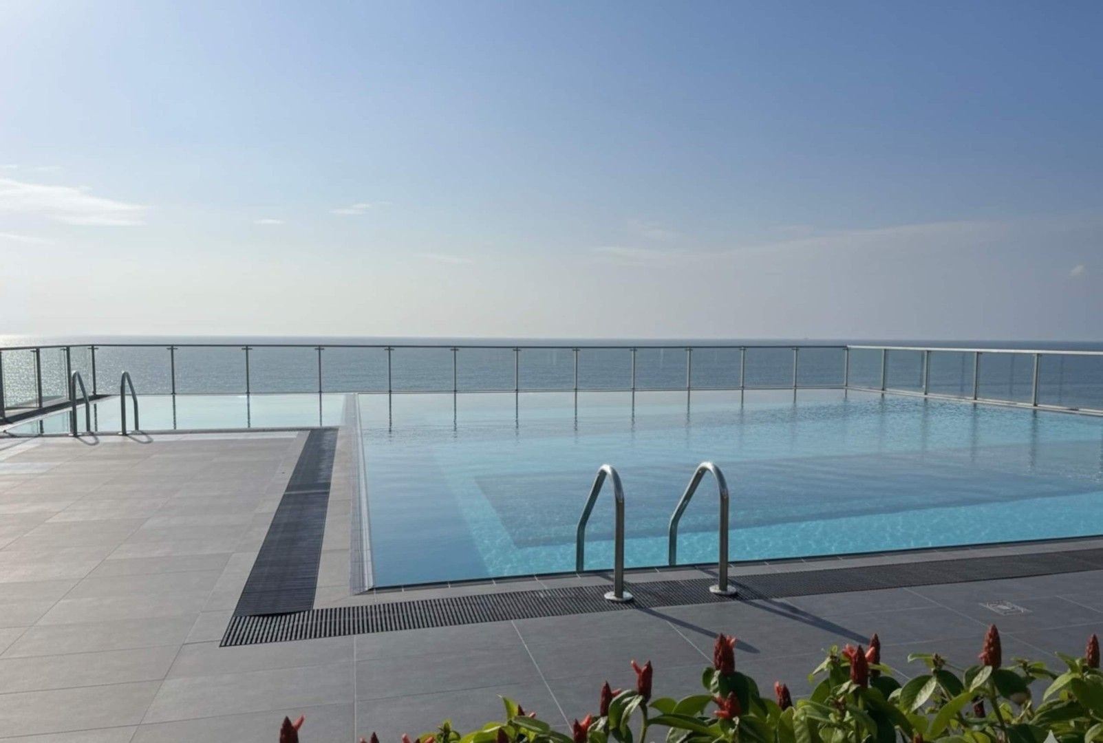 Una piscina azul cristalina con barandilla metálica, rodeada por una terraza con baldosas y vista al mar.