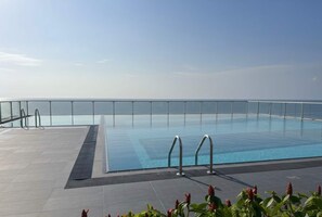 Ein klares, blaues Schwimmbecken mit Metallgeländer, umgeben von einer gefliesten Terrasse mit Blick aufs Meer.
