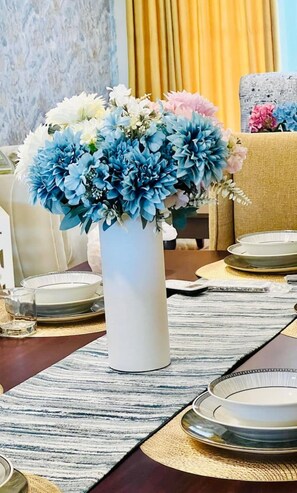 Ein Esstisch ist gedeckt mit weißen Tellern, einer hohen weißen Vase mit blauen und weißen Blumen und einer goldenen Tischmatte.