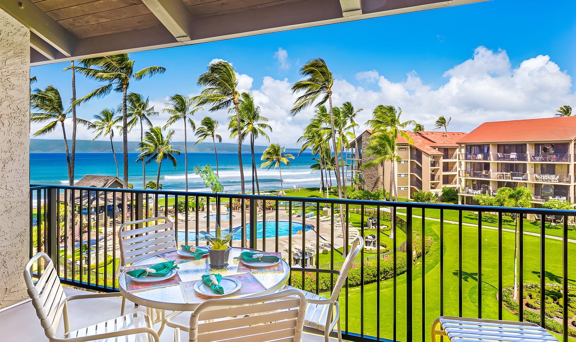 A balcony with a table set for dining, overlooking a pool and a beachfront hotel.