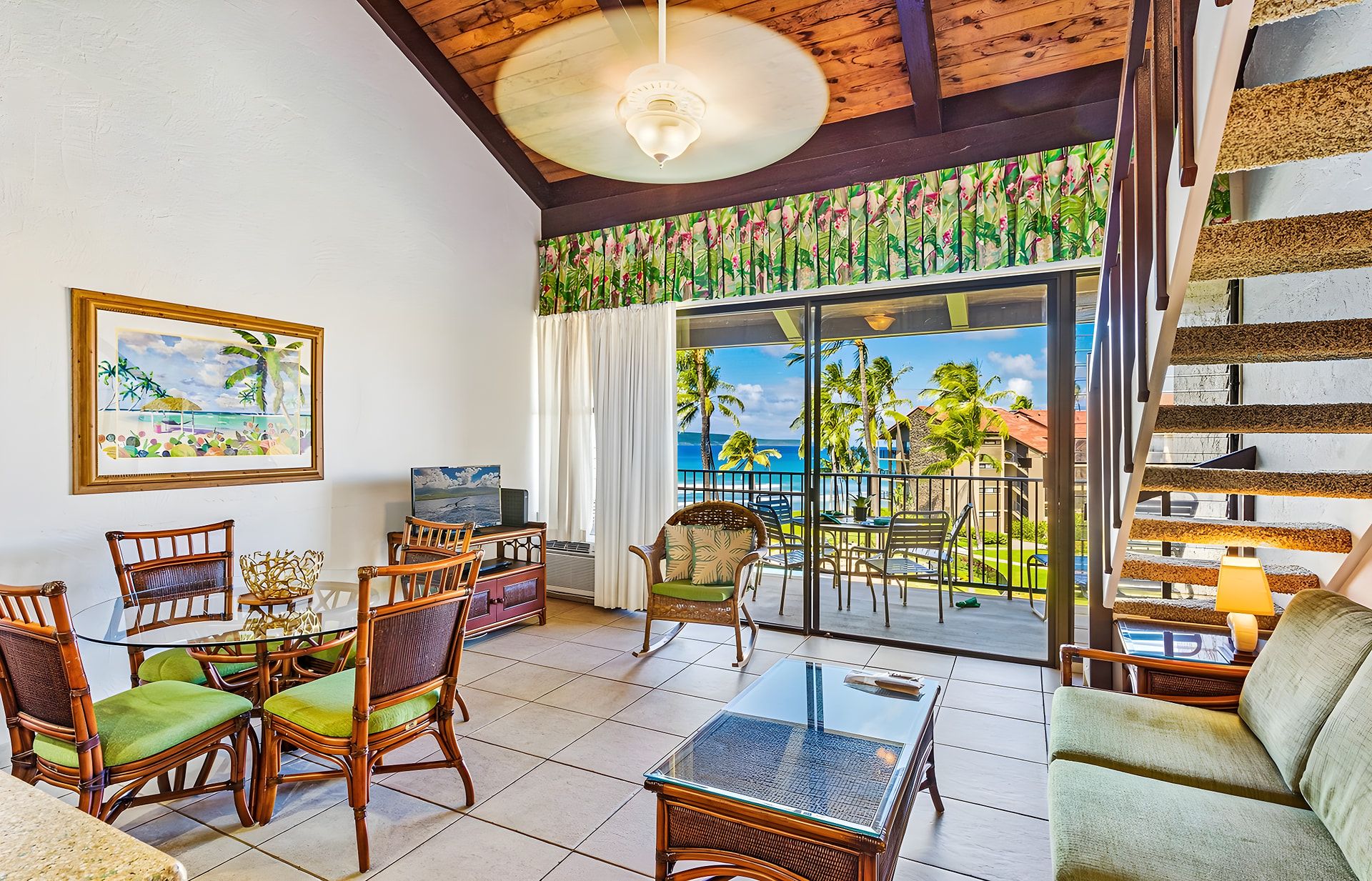 A spacious living area with a balcony overlooking a tropical beach, furnished with wicker chairs and a glass coffee table.