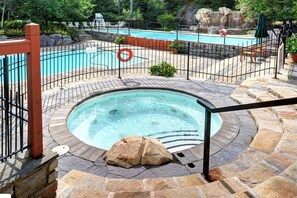 Ein Whirlpool mit Steinfassade, ein Pool mit schwarzem Zaun und Liegestühle.
