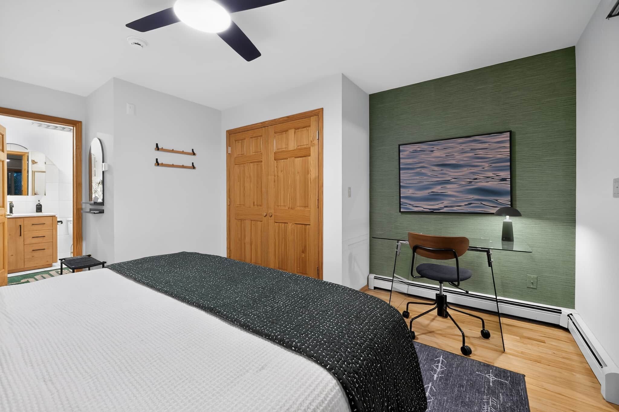 A bedroom with a bed, a wooden wardrobe, a desk with a chair, a ceiling fan, and a wall-mounted painting.