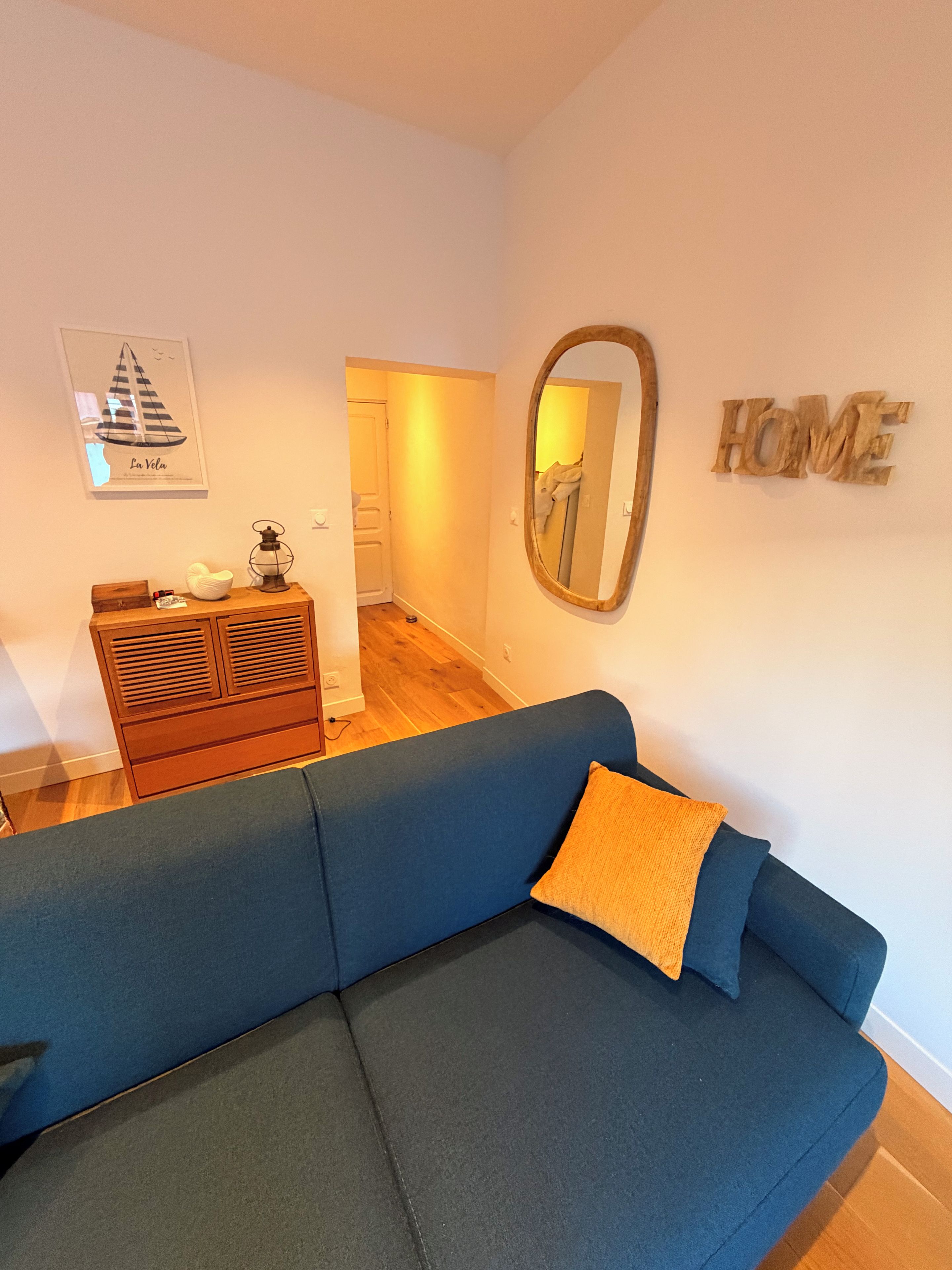 A living room with a blue sofa, a wooden dresser, a framed mirror, and a 'HOME' sign on the wall.