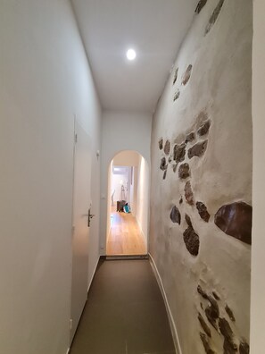 A hallway with a stone wall on one side and a door on the other. A wooden floor and a ceiling light are visible. - Renovated 3-Bedroom Apartment, 70 m²: Heart of the Old Port - Calanque Beach (La Ciotat)