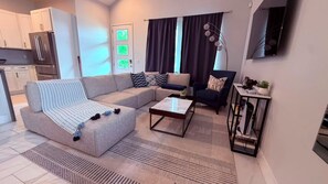 Una sala moderna con un sofá seccional, una mesita de centro y un sillón oscuro. Hay una zona de cocina con armarios blancos y un refrigerador.