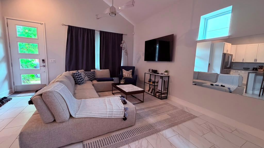 Uma sala de estar moderna com sofá, mesa de centro e televisão. Uma cozinha visível através de uma porta aberta.