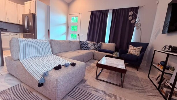 A modern living room with a sectional sofa, a coffee table, and a dark armchair. There is a kitchen area with cabinets and a refrigerator in the background. - Modern 3BR Haus in Lake Nona Medical City (Orlando, FL, 32827)